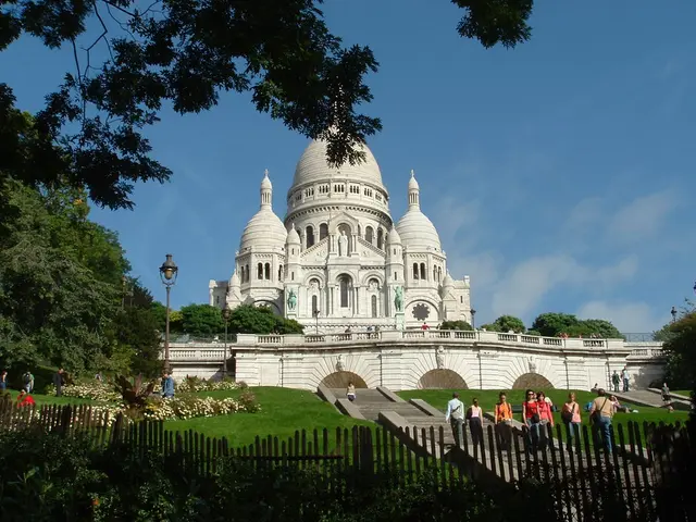 There are plants, fencing, grass, people, poles, white building and trees.